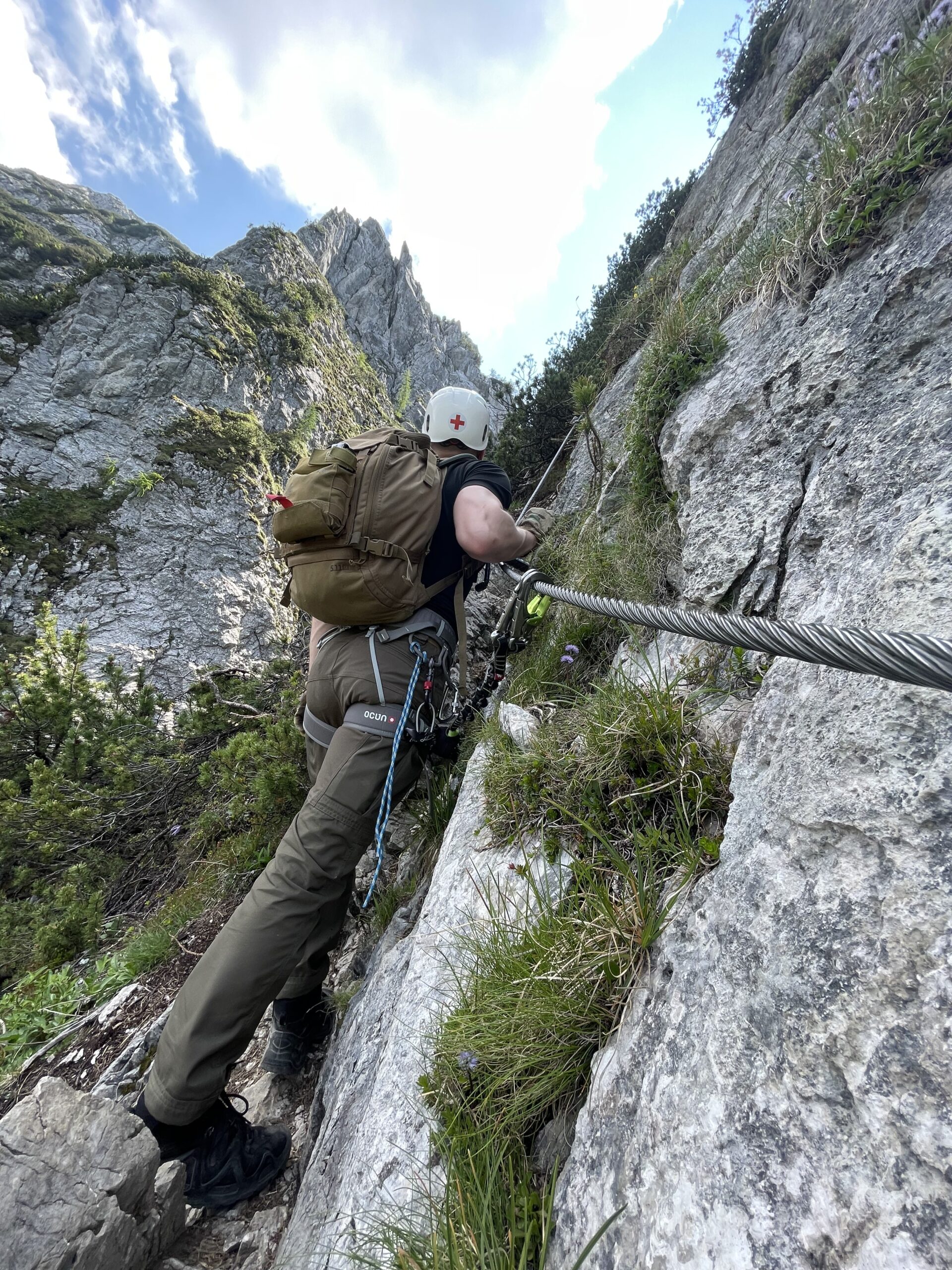 Helmeted climber with a backpack following a fixed steel cable through limestone terrain